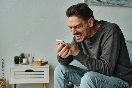 bearded man in grey jumper yelling at his smartphone while sitting on bed at home, emotional