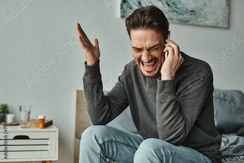angry bearded man in grey sweater talking on smartphone and gesturing in modern bedroom, argue