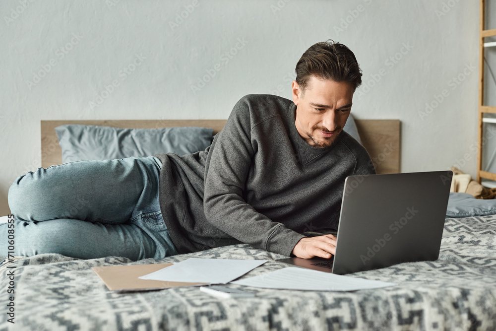focused and bearded man using laptop while working remotely from home in bedroom, documents focused and bearded man using laptop while working remotely from home in bedroom, documents