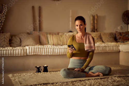 Portrait of a beautiful fit woman, sitting on the yoga mat, using a phone and earphones.