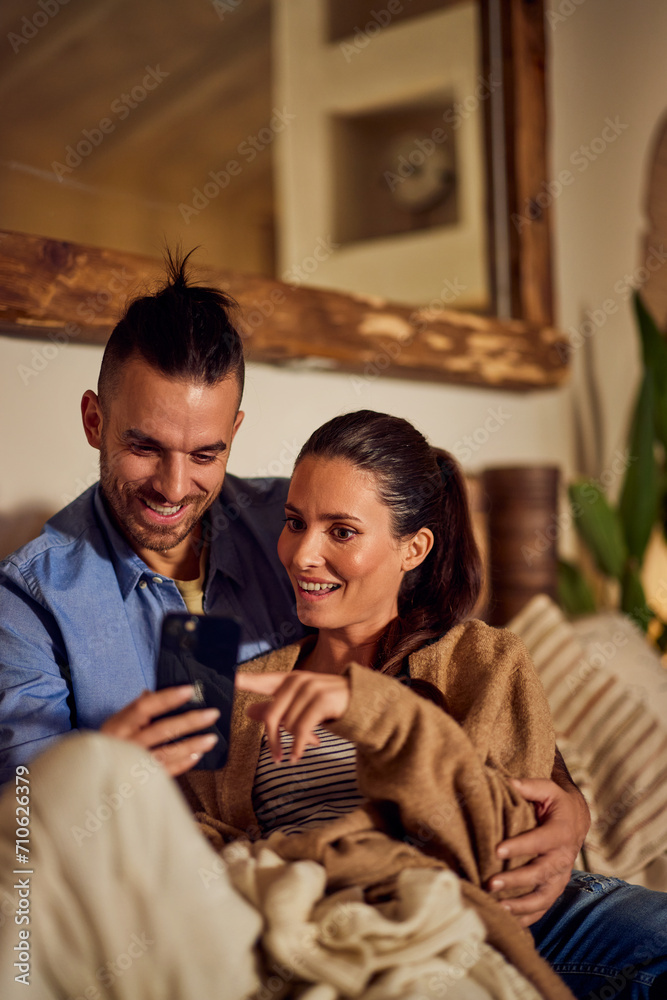 Smiling lovers enjoying the night at home, sitting on the couch, using a mobile phone.