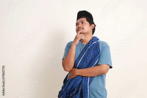 Confused young asian muslim boy standing while thinking or dreaming something. Isolated on white photo