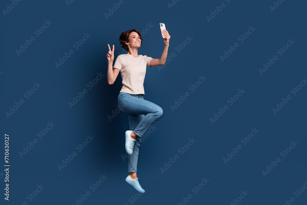 Full body photo of pretty young girl jump selfie device v-sign wear trendy white clothes isolated on dark blue color background Full body photo of pretty young girl jump selfie device v-sign wear trendy white clothes isolated on dark blue color background