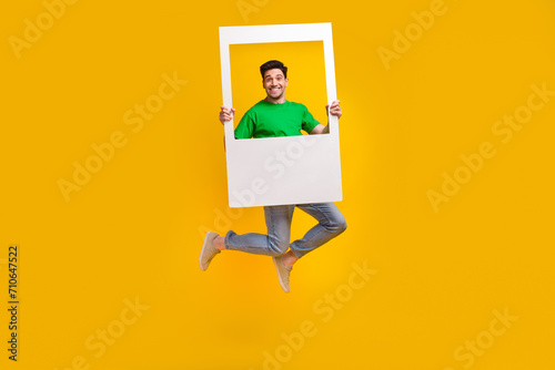 Full length photo of funny good mood pleasant man jumping with frame in hands make instant picture isolated on yellow color background