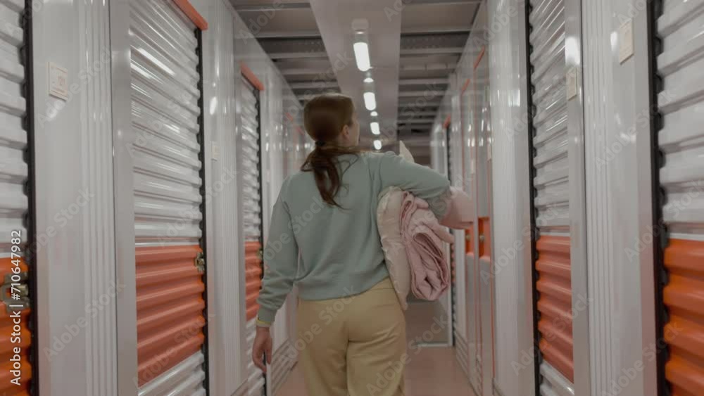 Rear view of woman with belongings enters in the self-storage facility unit. White and orange closed metal doors of storage warehouse. Moving, organizing, storage concept.