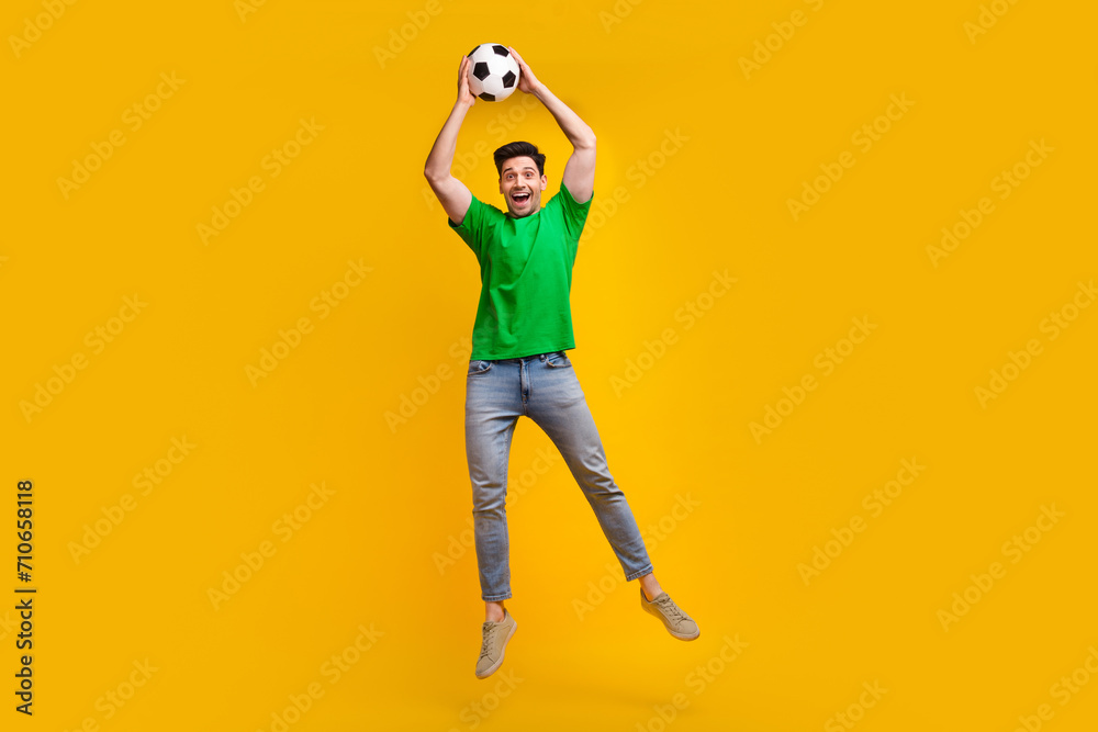 Fototapeta premium Full length photo of positive active man dressed green t-shirt jumping holding soccer ball over head isolated on yellow color background