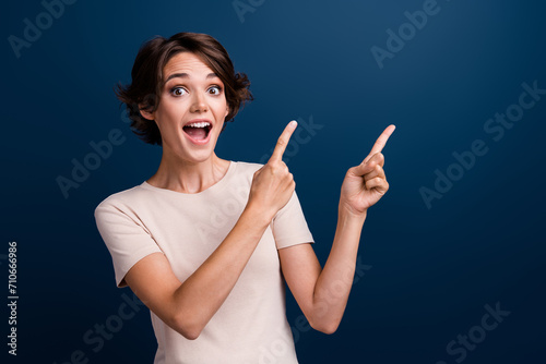 Portrait of funny amazed girl promoter directing fingers empty blank space cool news open mouth isolated dark blue color background photo