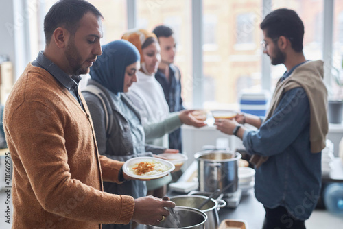 Side view portrait of adult Middle Eastern man receiving free hot meals at soup kitchen with people in line, copy space