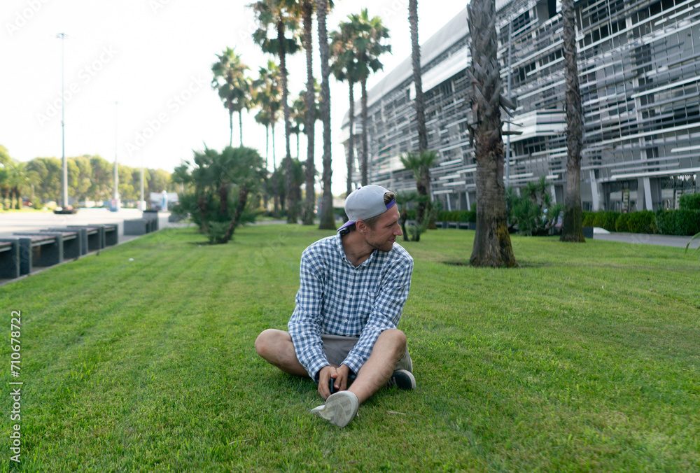 A man sits contentedly on a grassy lawn, enjoying a summer evening amidst tall palm trees and modern buildings, exuding a sense of peace and relaxation. A man sits contentedly on a grassy lawn, enjoying a summer evening amidst tall palm trees and modern buildings, exuding a sense of peace and relaxation.