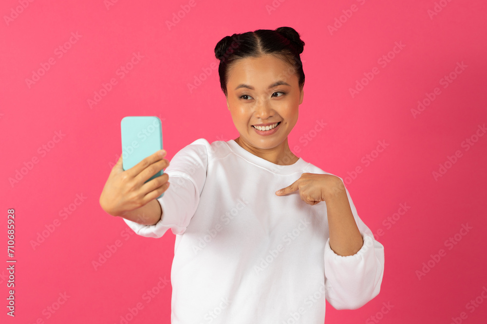 Happy young asian woman taking selfie on smartphone and pointing at herself Happy young asian woman taking selfie on smartphone and pointing at herself