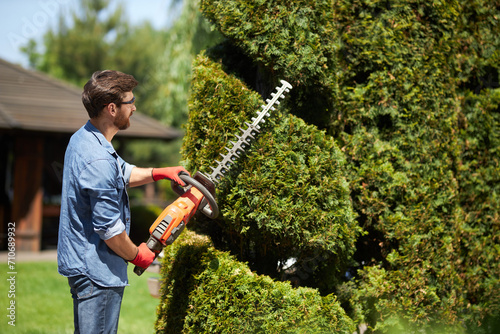 Skillful male landscaper shaping geometric ornament with hedge trimmer in topiary park. Side view of bearded professional gardener in gloves taking care of thuja with electric lopper. Topiary concept. photo