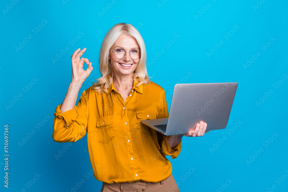 Photo of blonde lovely senior lady hold netbook show okey symbol wear trendy yellow silky garment isolated on blue color background
