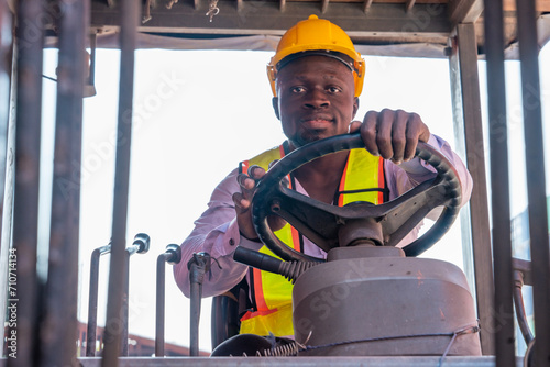 African American male worker wearing a hard hat sits and drives a forklift moving containers in a container yard.