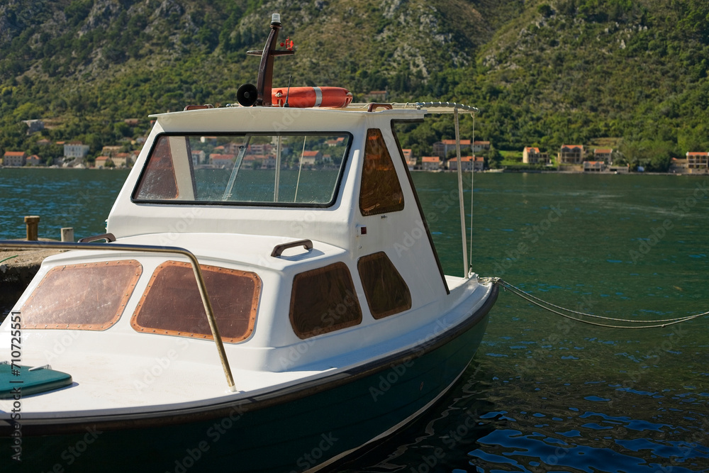 Naklejka premium A solitary white boat floats on the calm waters of Kotor Bay, the majestic backdrop of rugged mountains under a blue sky.
