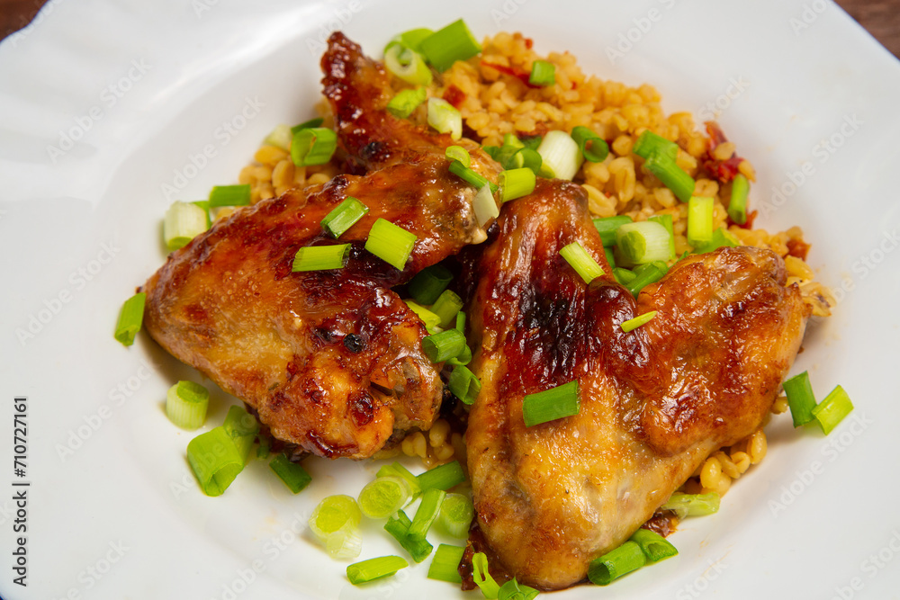 Chicken wings baked with bulgur, colored with green onions on a white plate, close-up