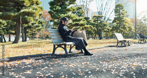 Japanese woman, bench and smartphone in park with relax, happiness and social media in sunshine in city. Female person, sitting and wellness with cellphone in town, streaming and digital technology