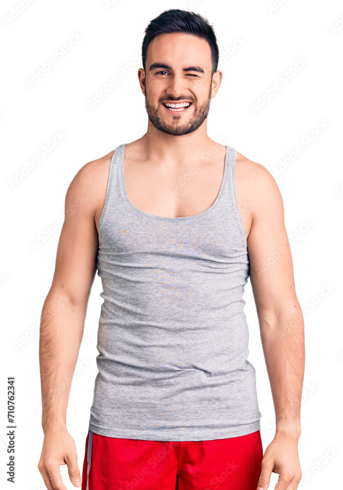 Young handsome man wearing swimwear and sleeveless t-shirt winking looking at the camera with sexy expression, cheerful and happy face. Young handsome man wearing swimwear and sleeveless t-shirt winking looking at the camera with sexy expression, cheerful and happy face.
