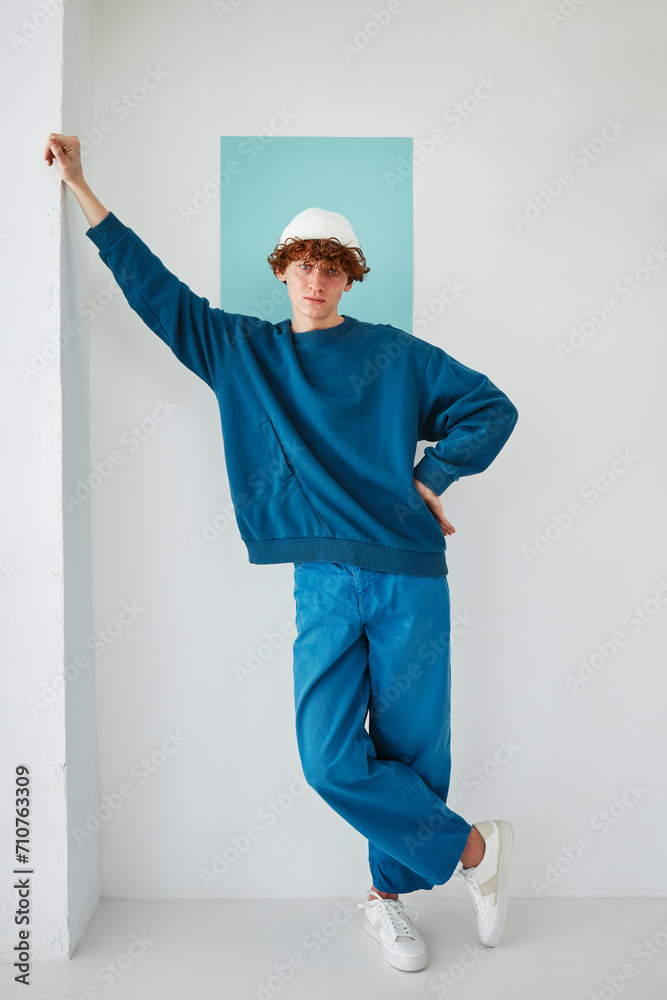 studio portrait on cyclorama of a young man with red curly hair in blue sweatshirt white beanie hat blue pants on blue paper background