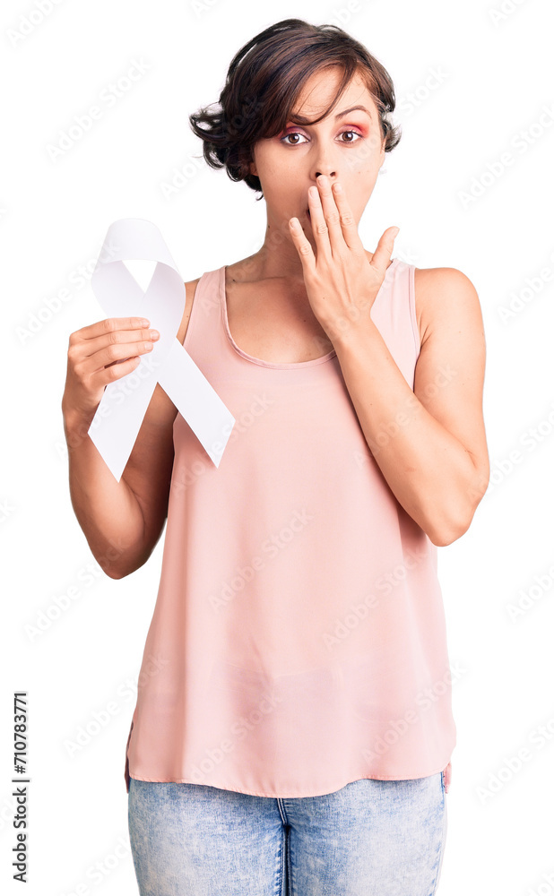 Beautiful young woman with short hair holding cancer awareness white ribbon covering mouth with hand, shocked and afraid for mistake. surprised expression Beautiful young woman with short hair holding cancer awareness white ribbon covering mouth with hand, shocked and afraid for mistake. surprised expression