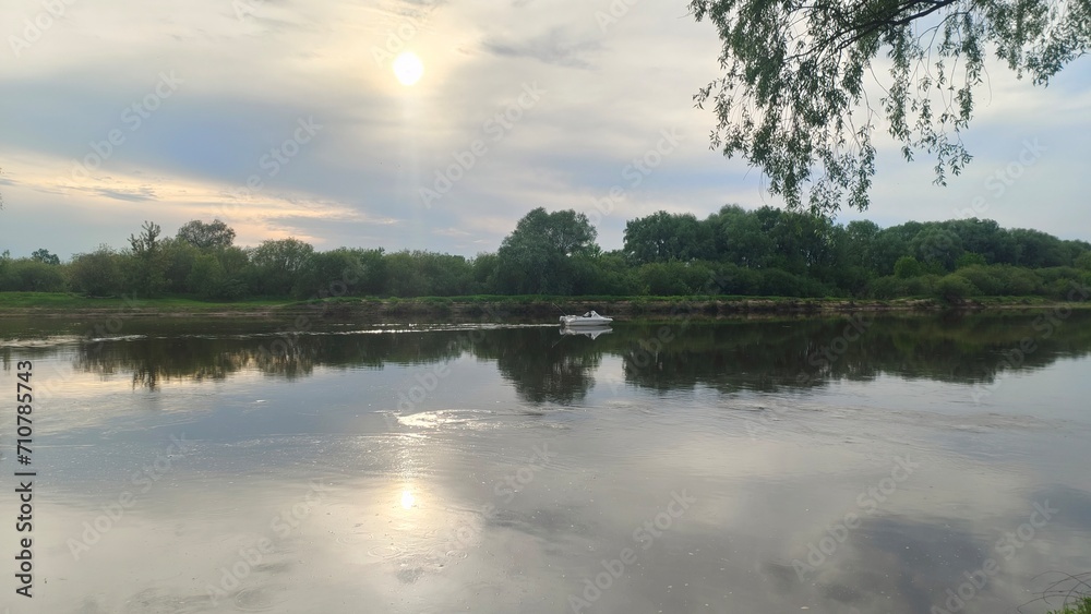 Before sunset, the summer sun is reflected in the water, overhung by willow branches. There is a motor plastic boat on the river and a fisherman trolling for predatory fish. Bushes and trees grow