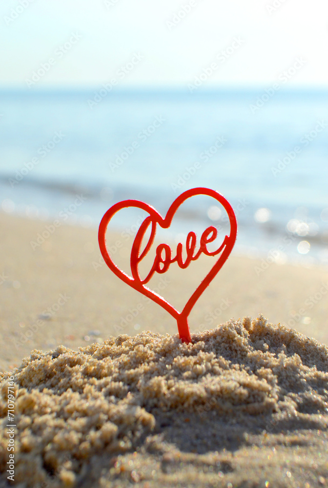 Plastic stick in shape of red heart and word Love in sand on beach seashore on sunny summer day close-up. Figures in shape of heart word Love on background sea waves. Love relationship romance concept Plastic stick in shape of red heart and word Love in sand on beach seashore on sunny summer day close-up. Figures in shape of heart word Love on background sea waves. Love relationship romance concept