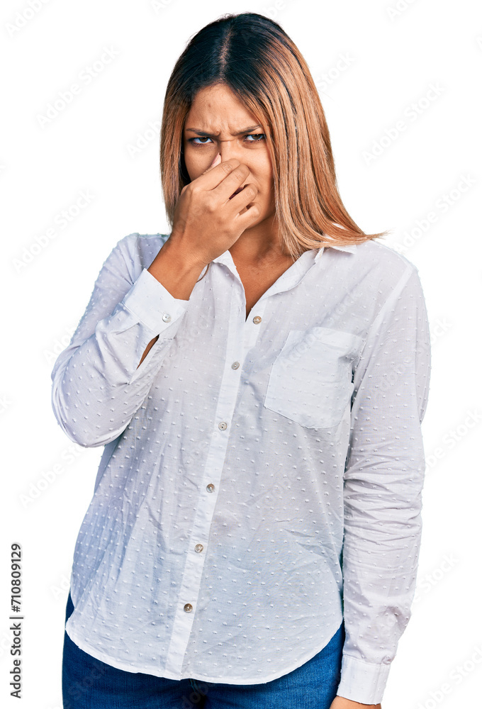 Young hispanic woman wearing casual white shirt smelling something stinky and disgusting, intolerable smell, holding breath with fingers on nose. bad smell