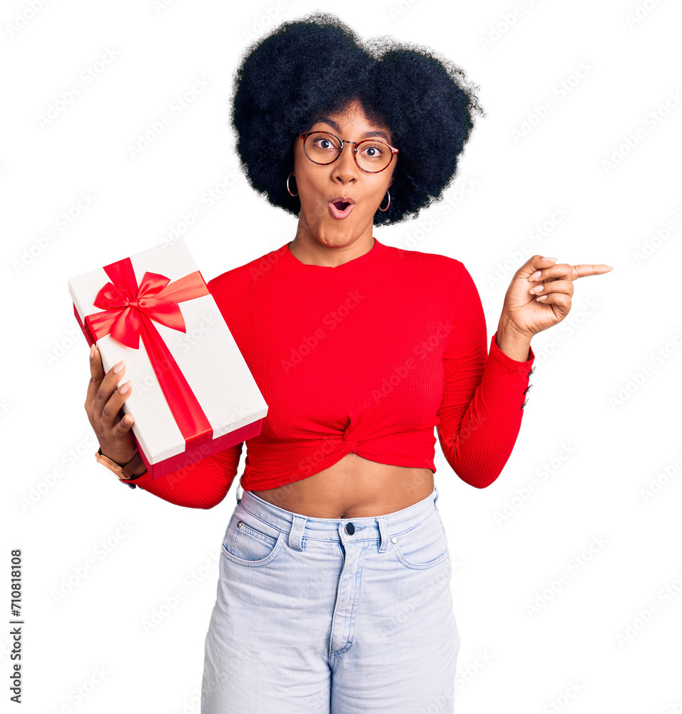 Young african american girl holding gift surprised pointing with finger to the side, open mouth amazed expression. Young african american girl holding gift surprised pointing with finger to the side, open mouth amazed expression.