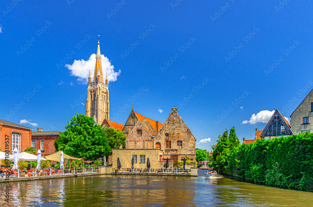 Fototapeta premium Bakkersrei water canal with tourist boat, Catholic Church of Our Lady Gothic style and Sint-Janshospitaal building in Brugge old town quarter, Bruges city historical centre, Flemish Region, Belgium