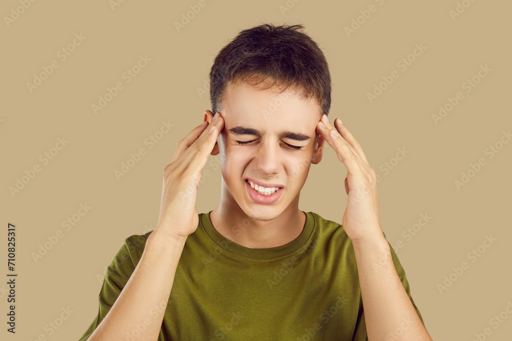 Close up portrait of a young student boy suffering from headache disease isolated on a studio beige background. Sick guy in casual clothes feel stressed, migraine or depression. Health care concept. Close up portrait of a young student boy suffering from headache disease isolated on a studio beige background. Sick guy in casual clothes feel stressed, migraine or depression. Health care concept.