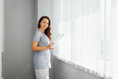 Beautiful smiling female doctor in medical uniform stands near the window in clinic. Young nurse is holding sheets of paper in her hands. Free Download Beautiful smiling female doctor in medical uniform stands near the window in clinic. Young nurse is holding sheets of paper in her hands. - GFXLoad