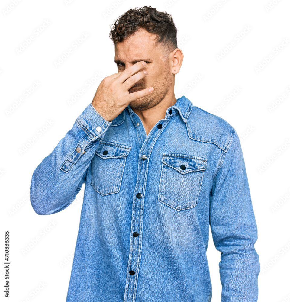Young hispanic man wearing casual denim jacket smelling something stinky and disgusting, intolerable smell, holding breath with fingers on nose. bad smell Young hispanic man wearing casual denim jacket smelling something stinky and disgusting, intolerable smell, holding breath with fingers on nose. bad smell