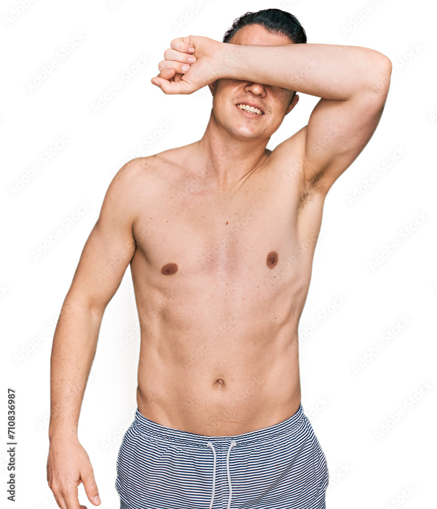 Handsome young man wearing swimwear shirtless covering eyes with arm smiling cheerful and funny. blind concept.