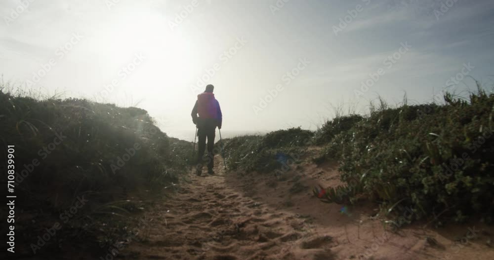 Camera follows solo male hiker walks along the trekking path towards the sun. Man silhouette with mountain poles reaching backpacking journey destination. Outdoor lifestyle. 