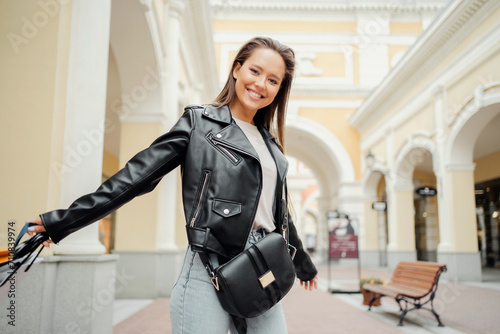 Joyful woman in leather jacket with a purse walking in a shopping arcade.