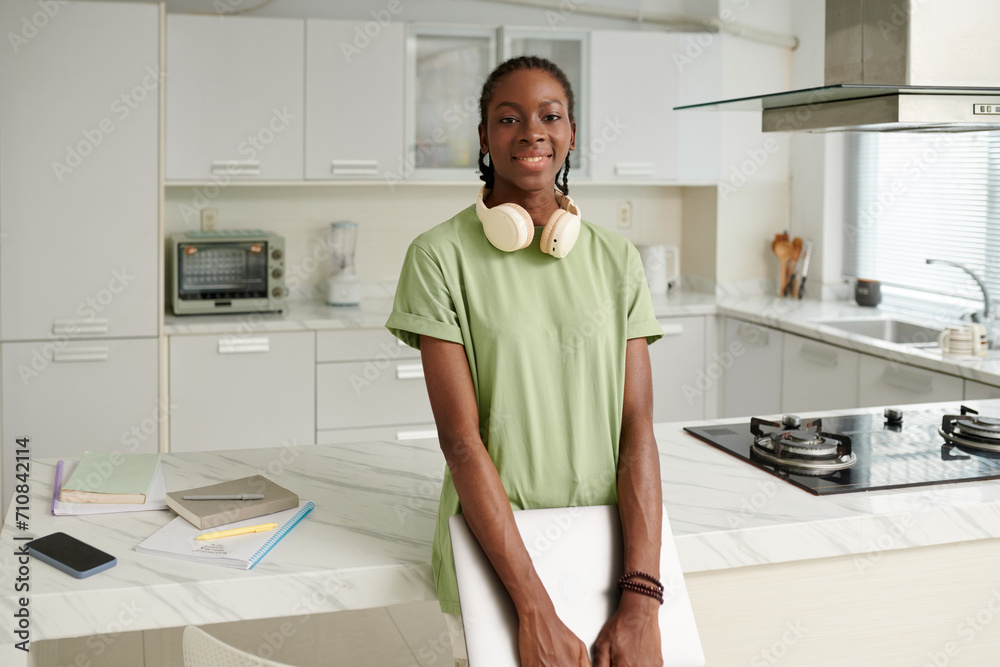 Cheerful Black girl with headphones and laptop standing at kitchen counter Cheerful Black girl with headphones and laptop standing at kitchen counter
