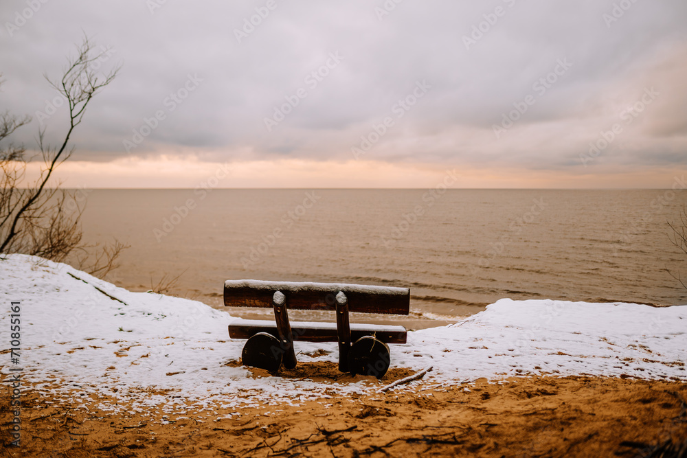 an empty wooden bench overlooking a calm sea, with snow on the ground and a cloudy sky above.