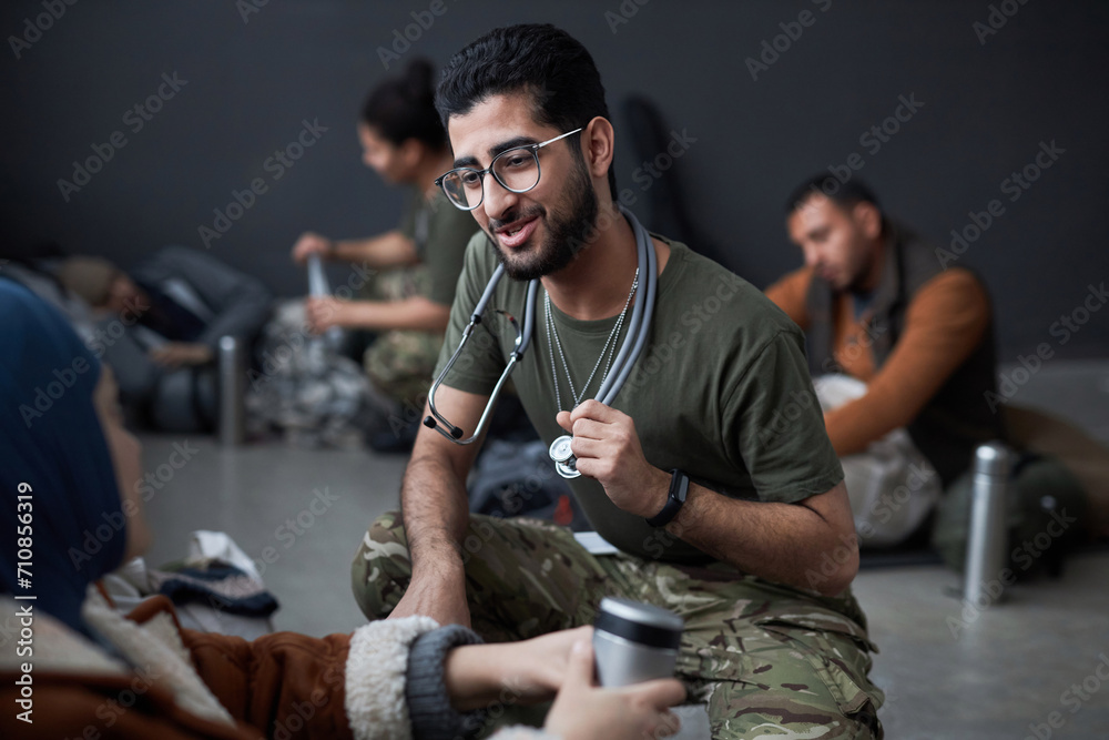 Portrait of smiling Middle Eastern man as military doctor talking to refugees and doing health examinations in shelter, copy space