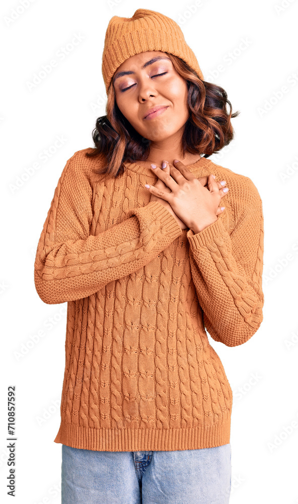 Young beautiful mixed race woman wearing wool sweater and winter hat smiling with hands on chest with closed eyes and grateful gesture on face. health concept.