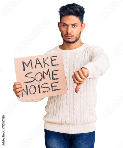 Handsome latin american young man holding make some noise banner with angry face, negative sign showing dislike with thumbs down, rejection concept