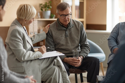 Portrait of senior man sharing mental health struggles with female psychologist in therapy session and holding glass of water, copy space