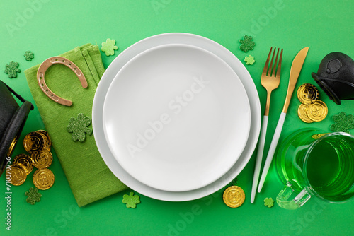 Setting the table for St. Paddy's celebration. Top view photo of plates, cutlery, napkin, leprechaun hat, pint of green beer, pots, coins, lucky horseshoe, clovers on green background