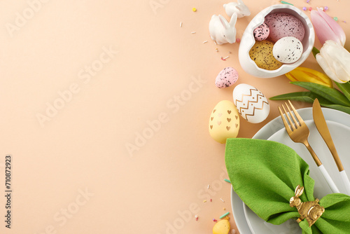 The Easter feast is presented with love and care. Top view shot of plates, cutlery, green napkin, painted eggs, fresh flowers, ceramic bunnies on beige background with congrats corner photo