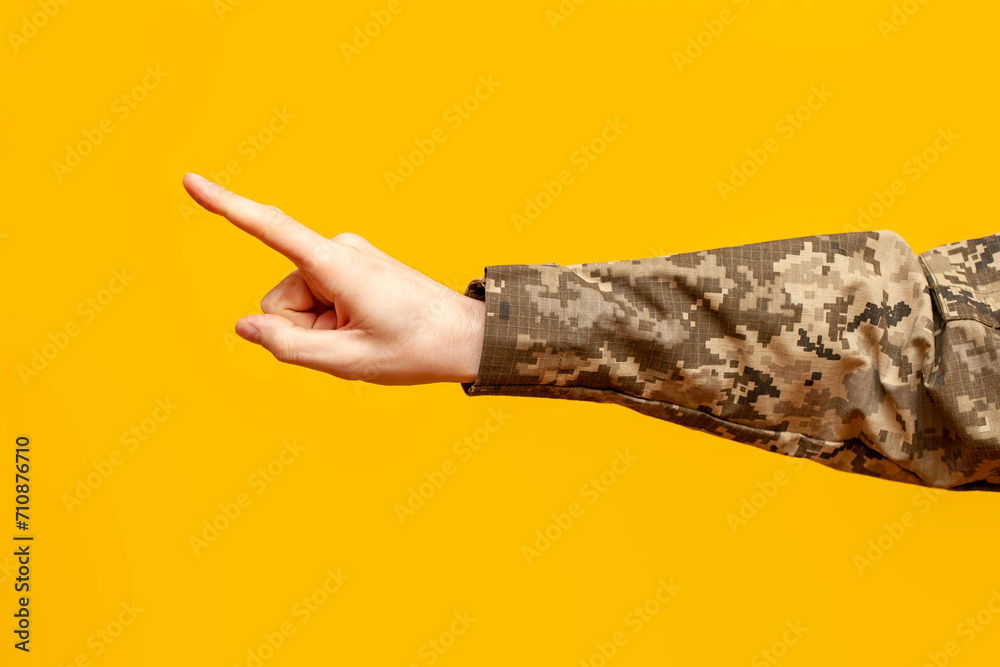 hand of a Ukrainian military man in camouflage uniform points with a finger and selects on a yellow isolated background, a male soldier points and presses, close-up - obrazy, fototapety, plakaty 