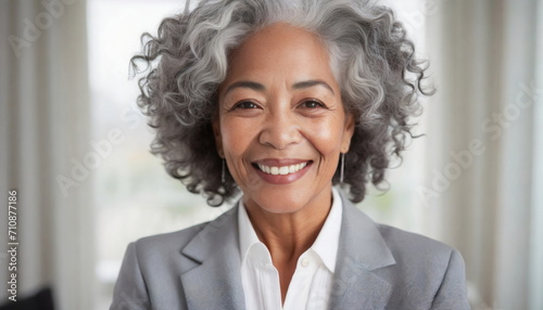 mature older woman with curly hairstyle, gray hair, wearing simple light gray suit and white shirt, smiling relaxed or proud