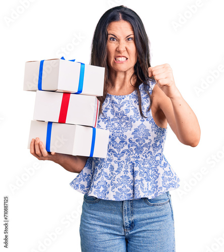 Young beautiful brunette woman holding birthday gift annoyed and frustrated shouting with anger, yelling crazy with anger and hand raised