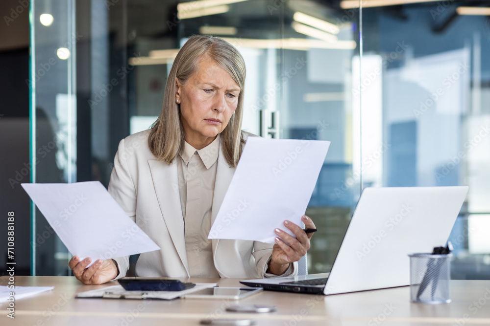 Worried senior businesswoman in suit working in office, sitting at desk, looking upset and analyzing documents. Worried senior businesswoman in suit working in office, sitting at desk, looking upset and analyzing documents.