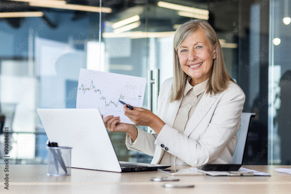 Portrait of a smiling senior successful business woman sitting in the office at the table, holding and showing documents and graphs to the camera.