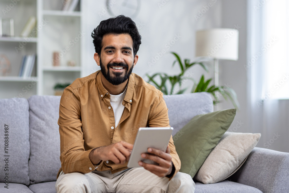 Portrait of a smiling young Indian man sitting on a sofa, holding a tablet and looking at the camera.