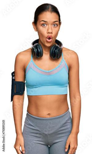 Beautiful hispanic woman wearing sportswear and headphones scared and amazed with open mouth for surprise, disbelief face