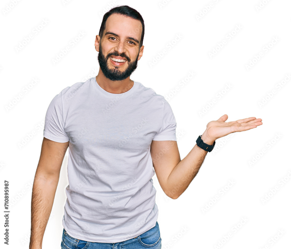 Young man with beard wearing casual white t shirt smiling cheerful presenting and pointing with palm of hand looking at the camera. Young man with beard wearing casual white t shirt smiling cheerful presenting and pointing with palm of hand looking at the camera.
